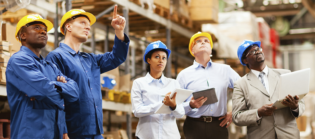 Two warehouse workers in blue shirts and hard hats point to an object above, as three individuals look on and take notes.